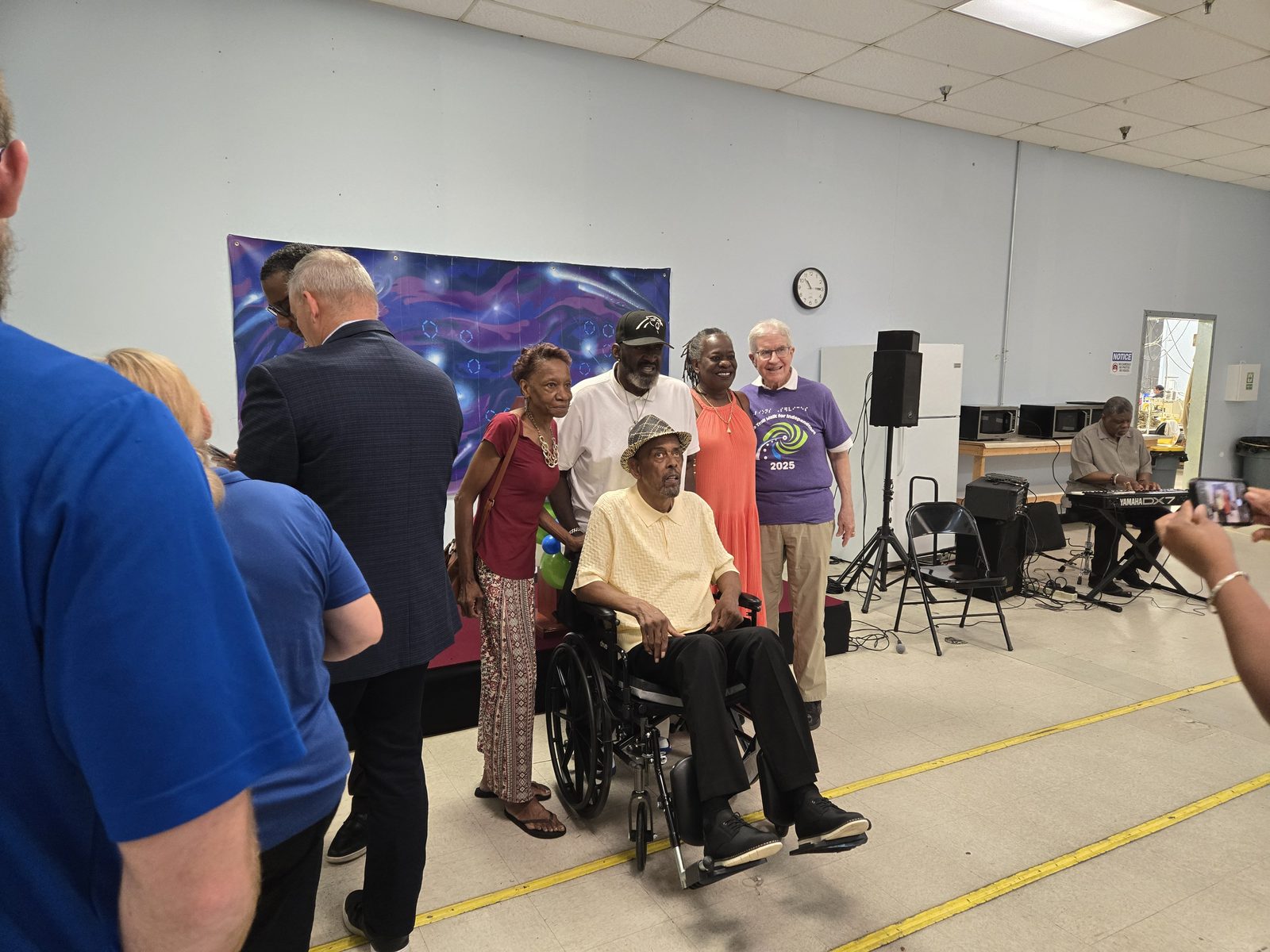 Guests gather indoors around an honored older gentleman seated in a wheelchair, posing together for a photograph in front of a starry backdrop, with a musician playing keyboard nearby.
