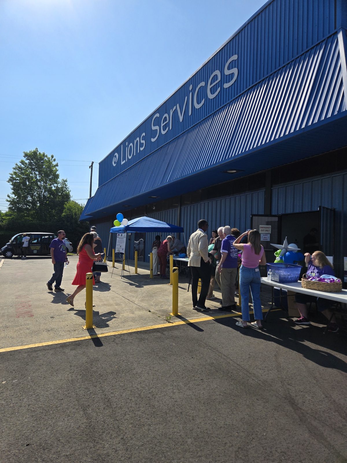 A crowd of guests arrives and checks in at welcome tables outside Lions Services, beneath blue and green balloons, on the morning of the Braille Trail reveal.