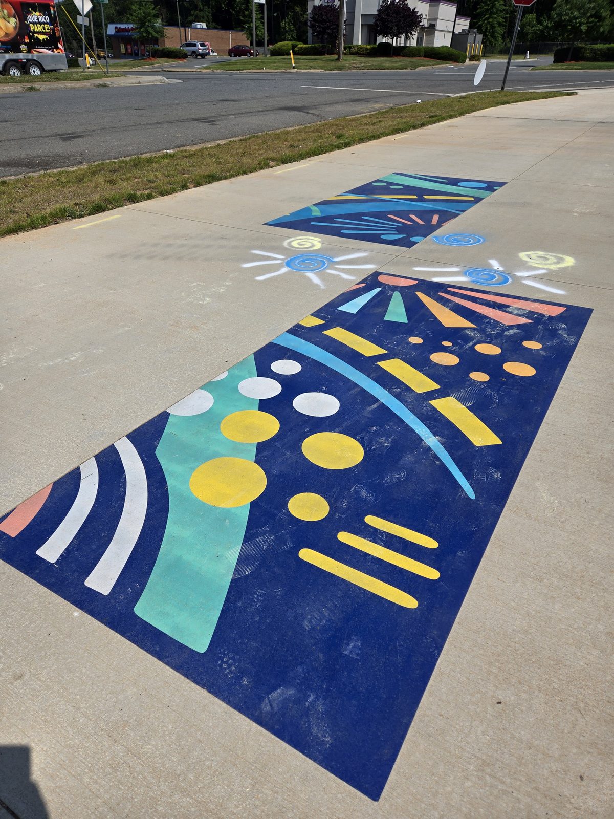 Two large, vibrant abstract murals painted directly onto the sidewalk along the trail, in bold blue, yellow, teal, white, and orange shapes, with chalk-drawn suns between them.