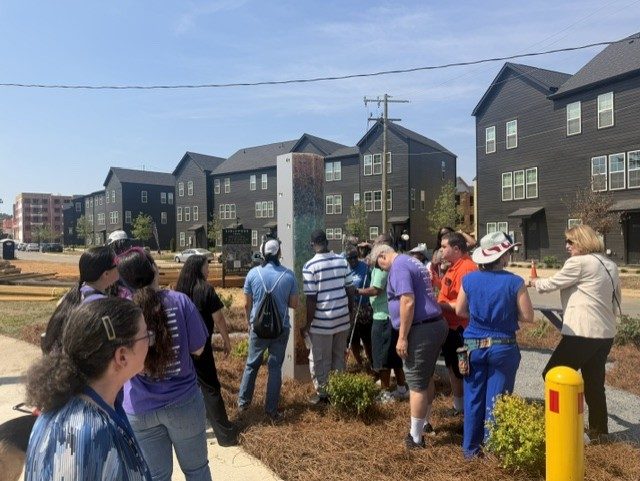 A group of visitors, many wearing purple Lions Services shirts, gather around a tall mirrored sculpture along the Braille Trail, with Charlotte townhomes in the background.