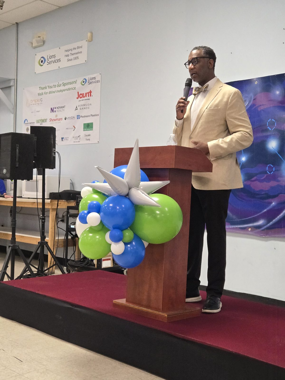 A speaker in a light blazer and bow tie addresses the audience from the balloon-decorated podium during the indoor reveal program.