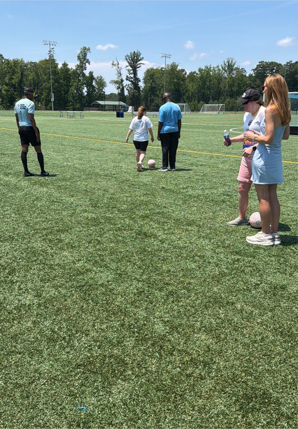 Participants and coaches gather on the turf during a blind soccer training drill, with goals and trees in the background.