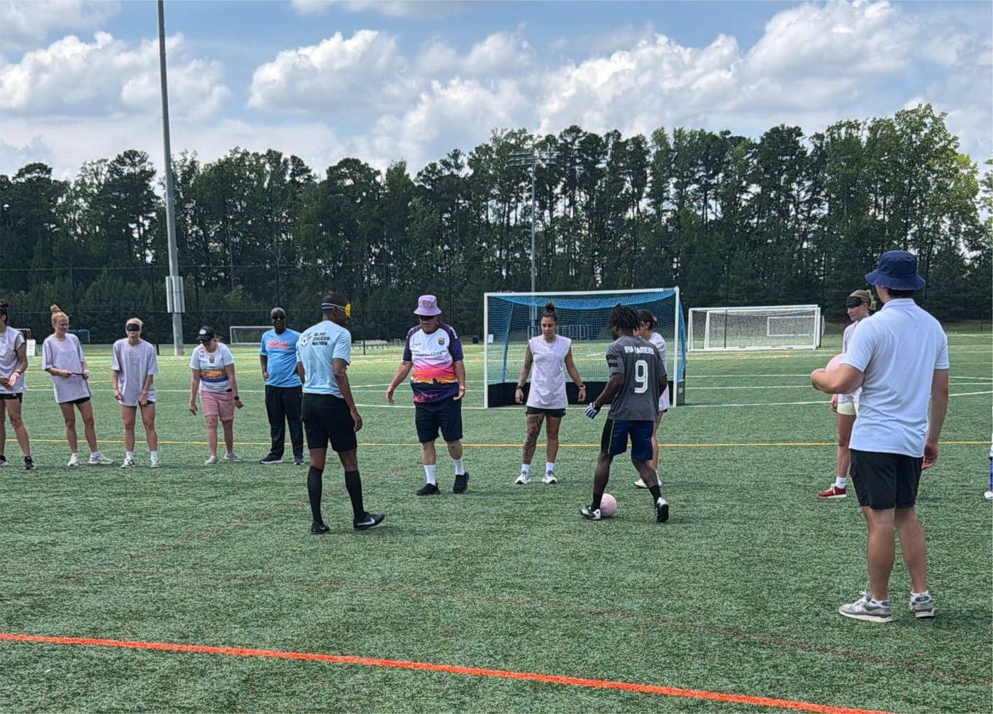 A large circle of participants listens to coaching on the turf field with a soccer goal and trees behind them.