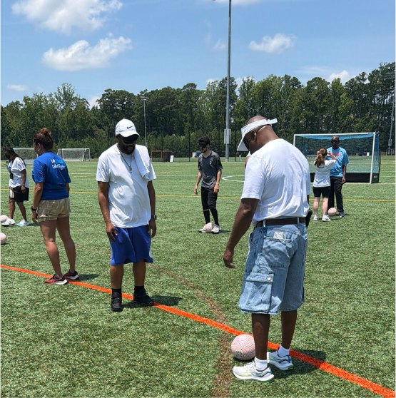Two athletes stand on the orange field line with a pink audible soccer ball between them during training.