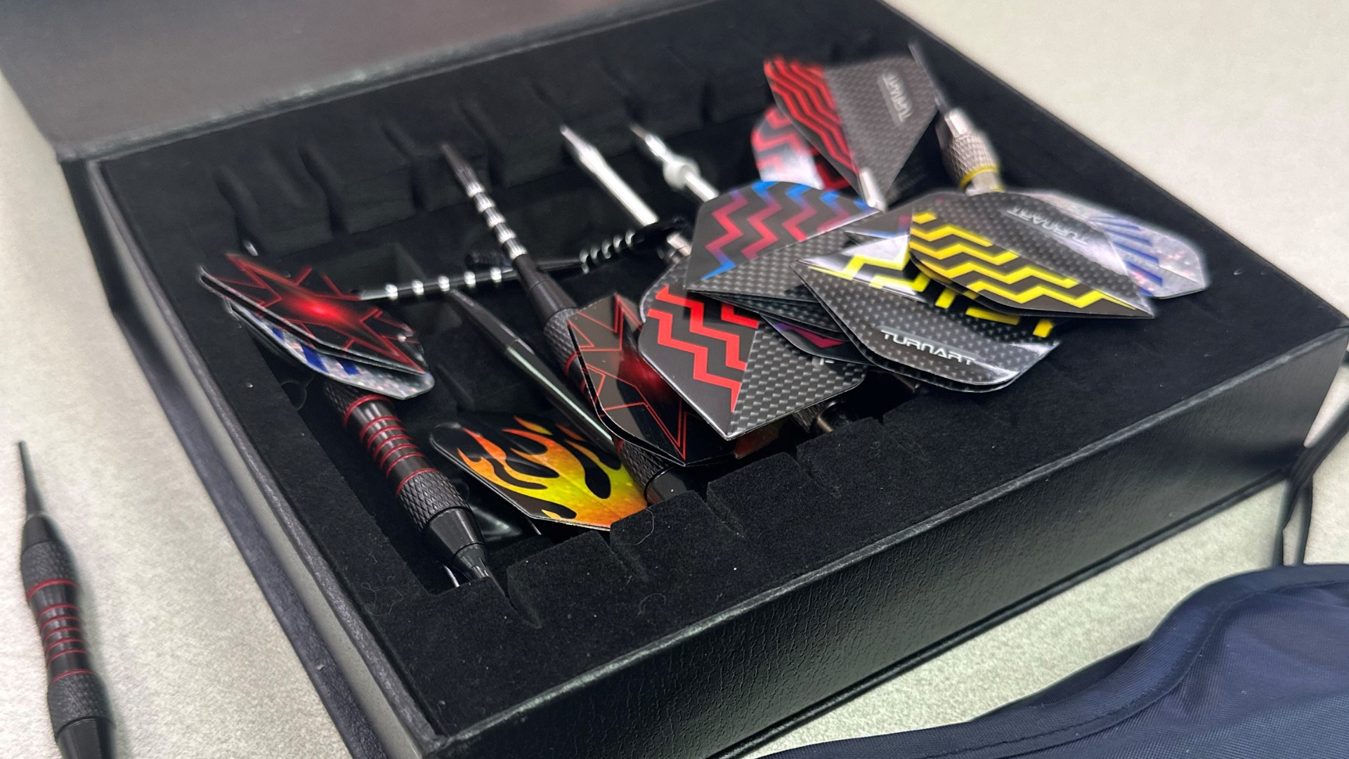 Image Shows a Box Full of Darts. The Flights have Flame Designs & Patterns. Many Darts inside a Bacl Box with Blindfold Tucked in Corner. Part of George's Collection of Darts.